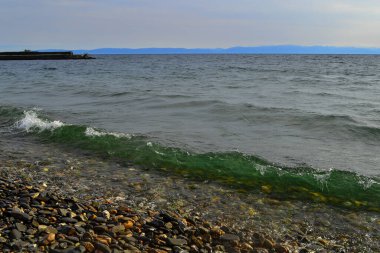 Yazın güneş ışığında parlak zümrüt yeşili baykal dalgaları, ufukta mavi dağlar, suda taşlar, sahilde deniz.
