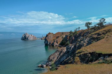 Şamanka Burnu berrak mavi Baykal Gölü 'nde, çimenli bozkırların arasında kozalaklı ağaçlar, kayalar, dağlar ve bulutların arka planında.