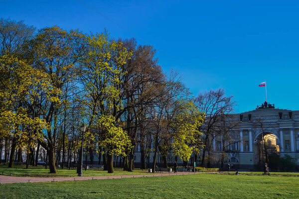 path in the park among the trees with yellow green foliage leaves in the light of the sun on the background of city architecture on the background of blue sky in spring