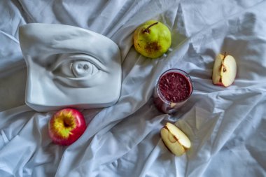 Olgun kırmızı elma, yeşil armut, buzlu meyve suyu ve David 'in göz heykeli beyaz kumaş örtüsü ve güneş ışığı altında kıvrımlar.