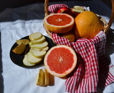 meyve sepeti, greyfurt elma yarımları kırmızı bir peçete üzerinde sepetle, armut dilimleri siyah tabakta beyaz bir masa örtüsüne karşı güneş ışığında