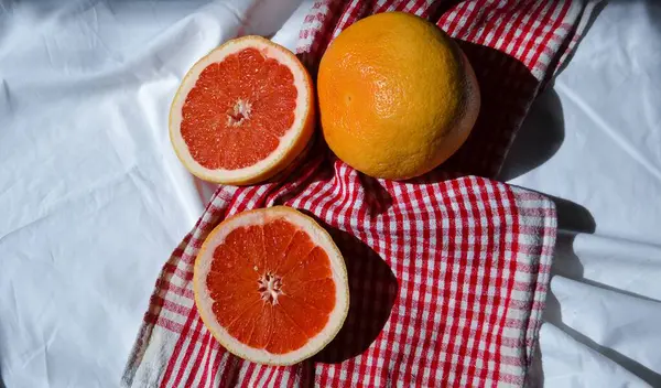 两半在白布和红色格子布上切下鲜红色的柚子 在阳光下折成褶皱 — 图库照片