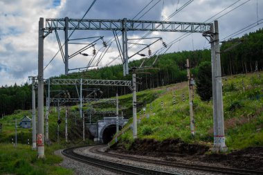 Demiryolu, yeşil dağ tepelerinde bir tüneli olan patika orman, tel sütunlar, gökyüzünde bulutlar