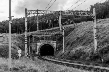 Demiryolu, tepelerde bir tünel, ağaçlar, tel sütunlar, gökyüzünde bulutlar, siyah ve beyaz