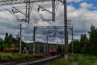 Demiryolu, kırmızı tren yeşil tepelerde ağaçlar, teller, gökyüzünde bulutlar