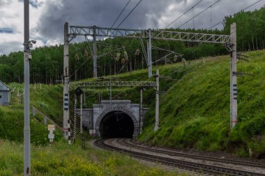 Demiryolu, yol yolu yeşil dağlarda bir tünel Ağaçlı tepeler, tel sütunlar, gökyüzünde bulutlar