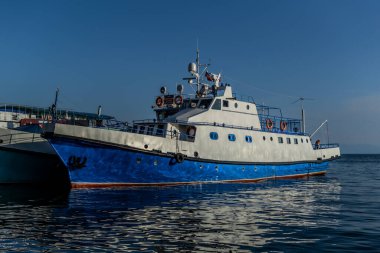 Baykal Gölü 'nün körfezindeki iskelede çizgili beyaz mavi gemiler. Güneş ışığı altında, suda yansımaları var.