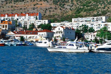 Beyaz renkli gemiler ve tekneler rıhtımlarda evlerin arasında suyla kaplı ve dağlarda kar maskesi inşa ediliyor, dalgalanmalar yansıyor.