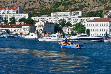Balaklava tatil köyü boyunca insanların yelken açtığı parlak mavi bir tekne. Evlerin ve binaların yakınındaki limanlarda beyaz gemiler arasında dalgalanmalar var.