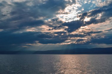 Akşamları parlak sarı pembe günbatımı. Gümüş gri bulutlar Baykal Gölü 'nde. Ufukta dağlar, güneş ışınları.