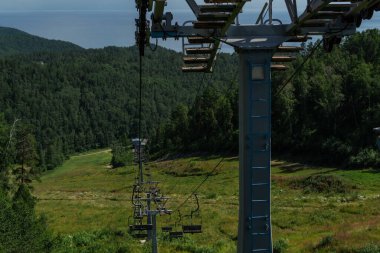 yeşil ağaçların arasında boş koltukları olan teleferik inşaatı, tepedeki ormanda çimen tarlası, beyaz örtülü güneşli yaz mavi gökyüzü, ufukta deniz.