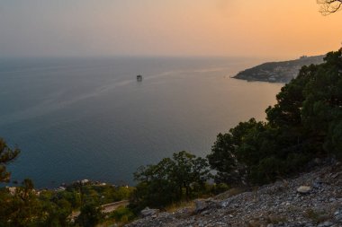 Karadeniz körfezi kıyısındaki tatil köyü manzarası dağlar ve kayalar arasında, mavi su, yaz, çalılar ve ağaçlar ön planda, günbatımı