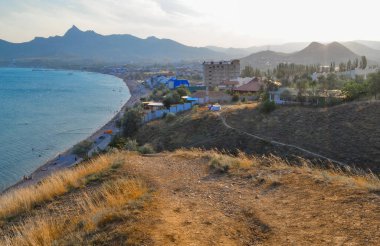 Karadeniz körfezi kıyısındaki tatil köyü manzarası dağlar ve kayalar arasında, mavi zümrüt suyu, yaz mevsimi, ön planda kuru sarı çimenler, gün batımı