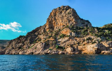 Kırmızı turuncu kaya kayalıkları mavi denizin kıyısında parlak güneşli bir günde bulutlu yaz gökyüzünde duruyor.