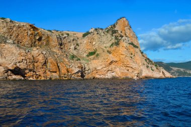 Kırmızı turuncu kayalıklar mavi denizin kıyısında parlak güneşli bir günde, bulutlu yaz gökyüzünde duruyor.