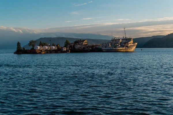 Küçük bir adada terk edilmiş eski beyaz gemiler. Mavi denizde ağaçlar, batan güneşin ışığında dağların ve bulutların ardında. Baykal Gölü