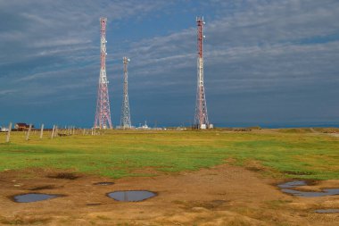 Sarı yeşil çimen, su birikintileri ve mavi gökyüzü ile yeşil alanda beyaz kırmızı renkli üç kulenin karşısında duruyor.