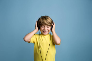 Eğlenceli genç çocuk kulaklıkla müzik dinleyerek ritim tutuyor. Sarı tişörtlü, gülümseyen sarışın çocuk.