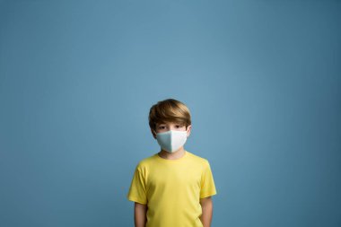 Covid 19 'u korumak için yüzünde tıbbi maske olan sevimli bir çocuğun portresi. Sarı tişörtlü sarışın çocuk.