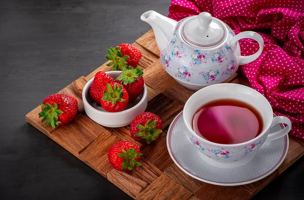 detail of a cup of strawberry tea accompanied by porcelain teapot and strawberries loose on a rustic wooden board with a crumpled towel blurred in the background