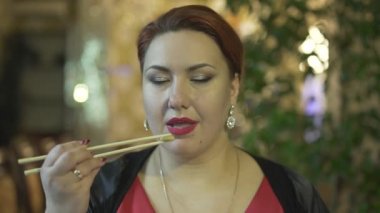 Çekici orta yaşlı esmer kadının yemek çubuklarıyla yemek yediği şık bir restoranda yemeğin tadını çıkardığı yakın plan.