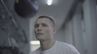 Kararlı genç erkek sporcu boksör boks salonunda ağır çekimde kum torbası yumrukluyor.