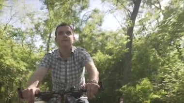 Mutlu, sakin, orta yaşlı, ekose gömlekli bir adam yaz güneşli bir günde, boş bir caddede yavaş bir bisiklet sürüyor.