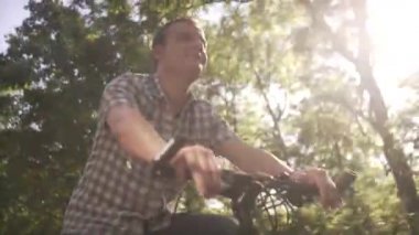 Düşük açılı mutlu, sakin, ekose gömlekli orta yaşlı bir adamın güneşli yaz gününde boş bir sokakta yavaş bir şehir bisikleti sürüşü.