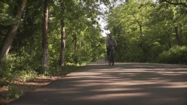 夏日阳光明媚 快乐中年男子骑着自行车在空旷的自行车道上走过绿树成荫的公园 拍摄角度很低 — 图库视频影像