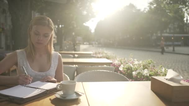 ブロンド若いです女性で白いドレスで外のカフェテラステーブル書き込み思考で個人ジャーナル日記離れて見て — ストック動画
