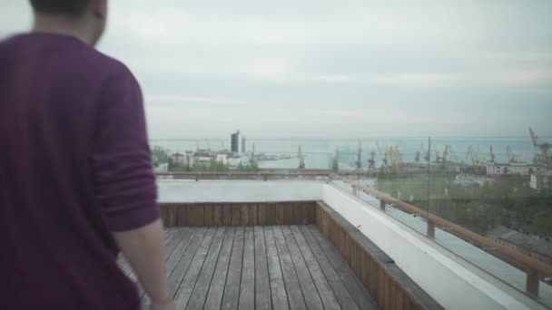 暗い髪の男カジュアル服でジャーナルで手を歩いて木の屋上に曇りの日に港の風景を見て — ストック動画