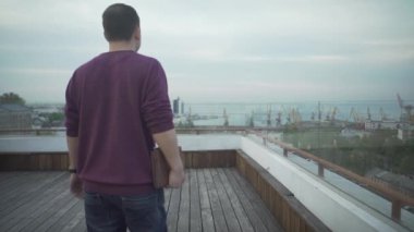 Elinde günlük tutan koyu renk saçlı bir adam bulutlu bir günde ahşap çatıda yürüyen deniz manzarasına bakıyor.