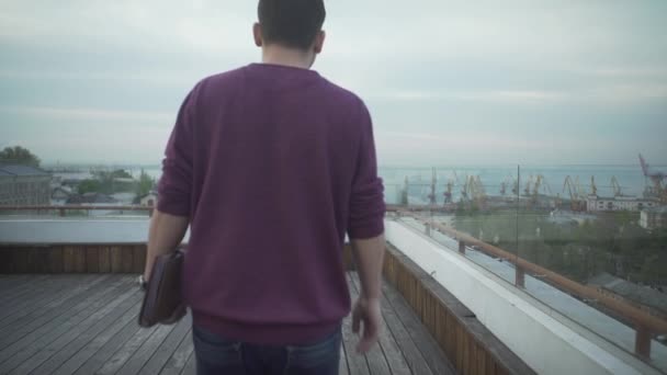 暗い髪の男でカジュアルな服で日記を手に海の港を見て曇りの日に木の屋上を歩く — ストック動画