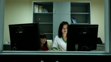 Küçük tatlı kız, genç, çekici, siyah saçlı kadına gülümsüyor. İş yerindeki iki bilgisayar ekranının arkasındaki doktor.