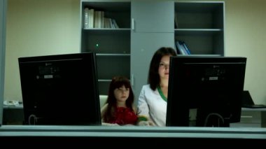 Küçük tatlı kız, iş yerindeki iki bilgisayar ekranının arkasındaki güzel siyah saçlı kadına gülümsüyor.