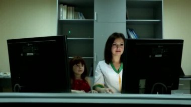 Şirin küçük kız, iş yerindeki iki bilgisayar ekranının arkasındaki güzel siyah saçlı kadına gülümsüyor.