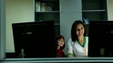 Güzel genç bayan doktor iki bilgisayar ekranının arkasında oturmuş gülümsüyor mutlu, heyecanlı küçük kız parmağını gösteriyor.
