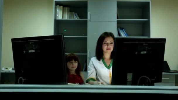 Kleines nettes Mädchen lächelt junge schöne dunkelhaarige Ärztin hinter zwei Computermonitoren im Büro an