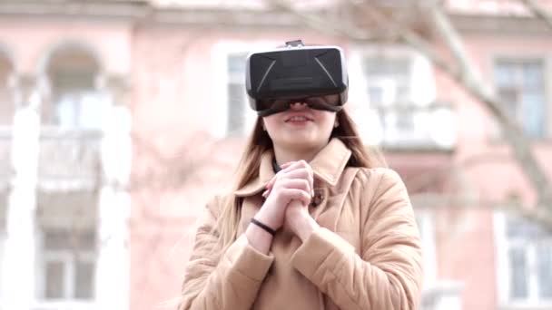 年轻快乐的女孩戴着虚拟现实耳机眼镜 穿着米色外套在外面的街上玩得很开心 — 图库视频影像