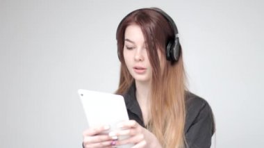 Kulaklıklı güzel genç bir kadın beyaz arka planda tabletle oynuyor.