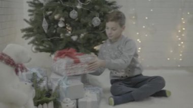 Heyecanlı küçük çocuk Noel hediyesi kutusunu dekore edilmiş yeni yıl ağacı şenlikli atmosfer odasında açıyor.