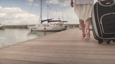 Çekici, formda bir kadın ağır bavullarla geri dönüyor. Yat limanındaki ahşap köprüde yürüyor.