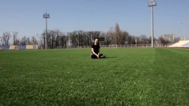 Çekici genç, aktif, fit bir kadın büyük stadyum yeşil çimenlerde oturmuş gülüyor ve güneşli bahar gününün keyfini çıkarıyor.