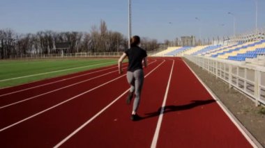 Genç, çekici, atletik bir kadın. Stadyumda ağır çekimde koşuyor. Güneşli bir bahar gününde açık havada egzersiz yapıyor.