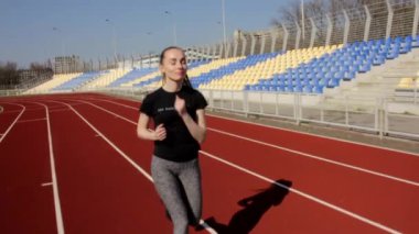 Genç atletik çekici kadın ağır çekimde koşuyor, güneşli bahar gününde açık havada büyük bir stadyum idmanı yapıyor.