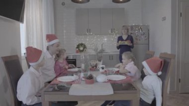 Heyecanlı mutlu dört çocuk ailesi yeni yılı kutluyor mutfak masasında baba oğul gülümseyen anne yemek servisi yapıyor.
