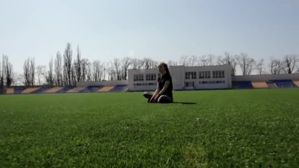 若い魅力的なアクティブフィット女性が巨大なスタジアムの緑の芝生の上に座って静かに暖かい晴れた春の日を楽しんで瞑想 — ストック動画