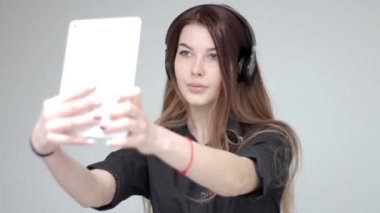 Kulaklıklı genç bayan beyaz tabletle selfie çekerken poz veriyor.