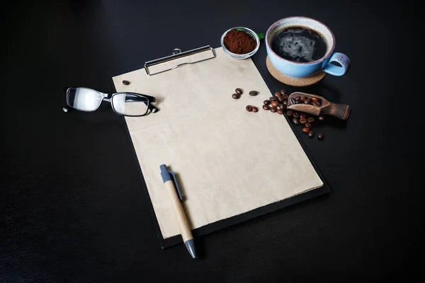 Menu and coffee. Clipboard with blank kraft letterhead, pen, coffee cup, coffee beans, pen, glasses and ground powder on black table background.