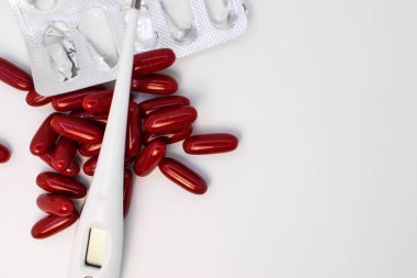 Sol tarafta beyaz bir arka planda kırmızı kapsüller arasında beyaz bir elektronik termometre var. Tedavi ve sağlığınıza dikkat etme kavramı